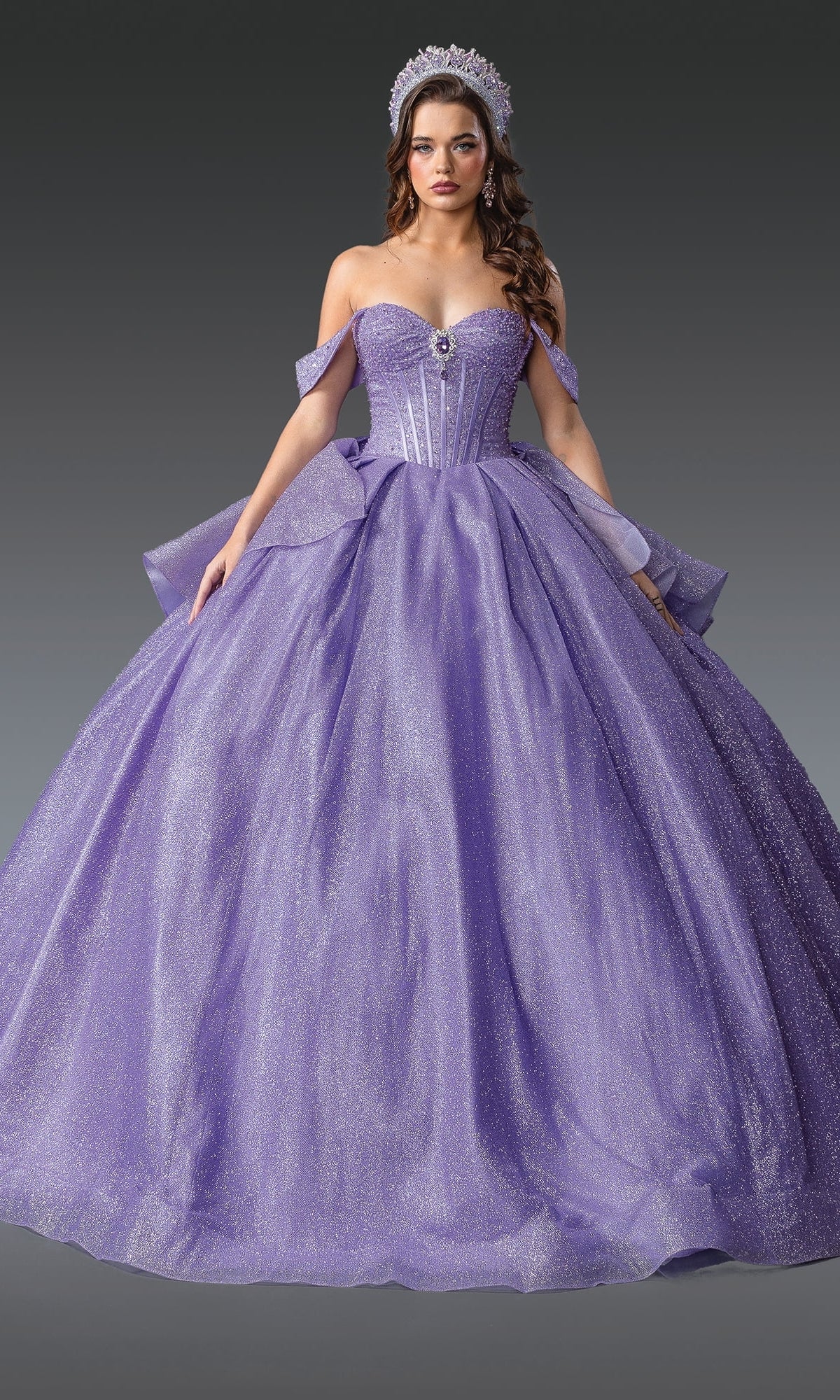 A woman wearing the Quinceanera Dress 1937 By Dancing Queen, a sparkling lavender ball gown with off-the-shoulder straps and a full skirt, plus a tiara, stands against a plain gray background.